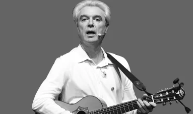 David Byrne performing live, playing an acoustic guitar, captured in a striking black and white photo.