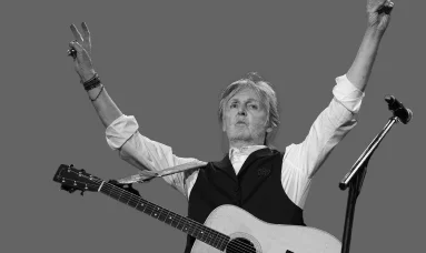 Paul McCartney raising his arms in a triumphant gesture, with a guitar strapped across his body, captured in black and white.