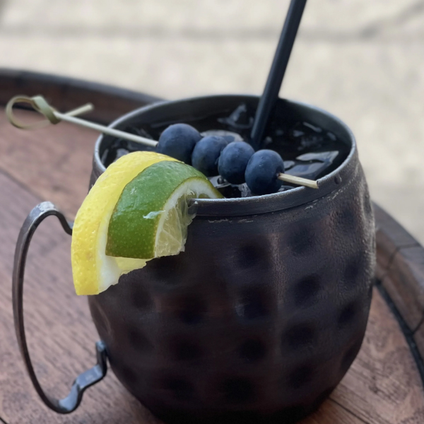 Close-up of a stylish black cocktail mug filled with ice, garnished with blueberries and a lime wedge, served on a wooden table.